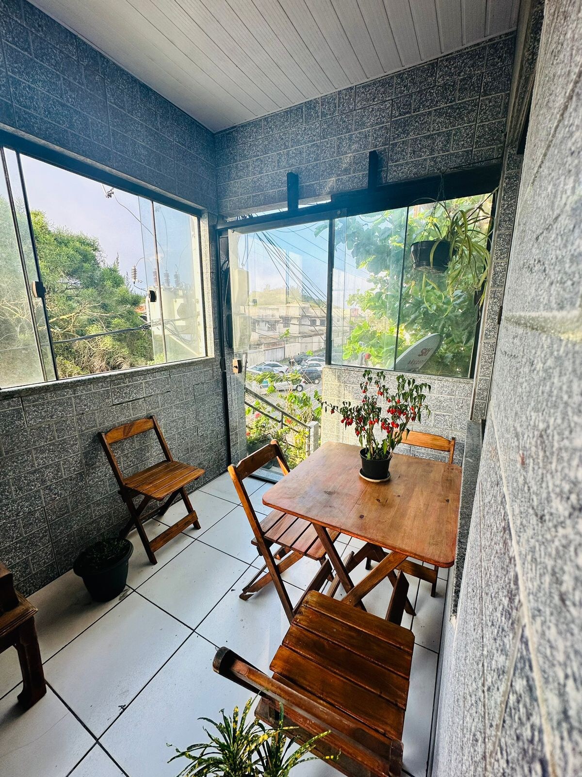 A shared balcony area is depicted, featuring a wooden table surrounded by four folding chairs. Natural light enters through large windows, highlighting plants that add a touch of greenery to the space. Views of the surrounding area are partially visible outside.