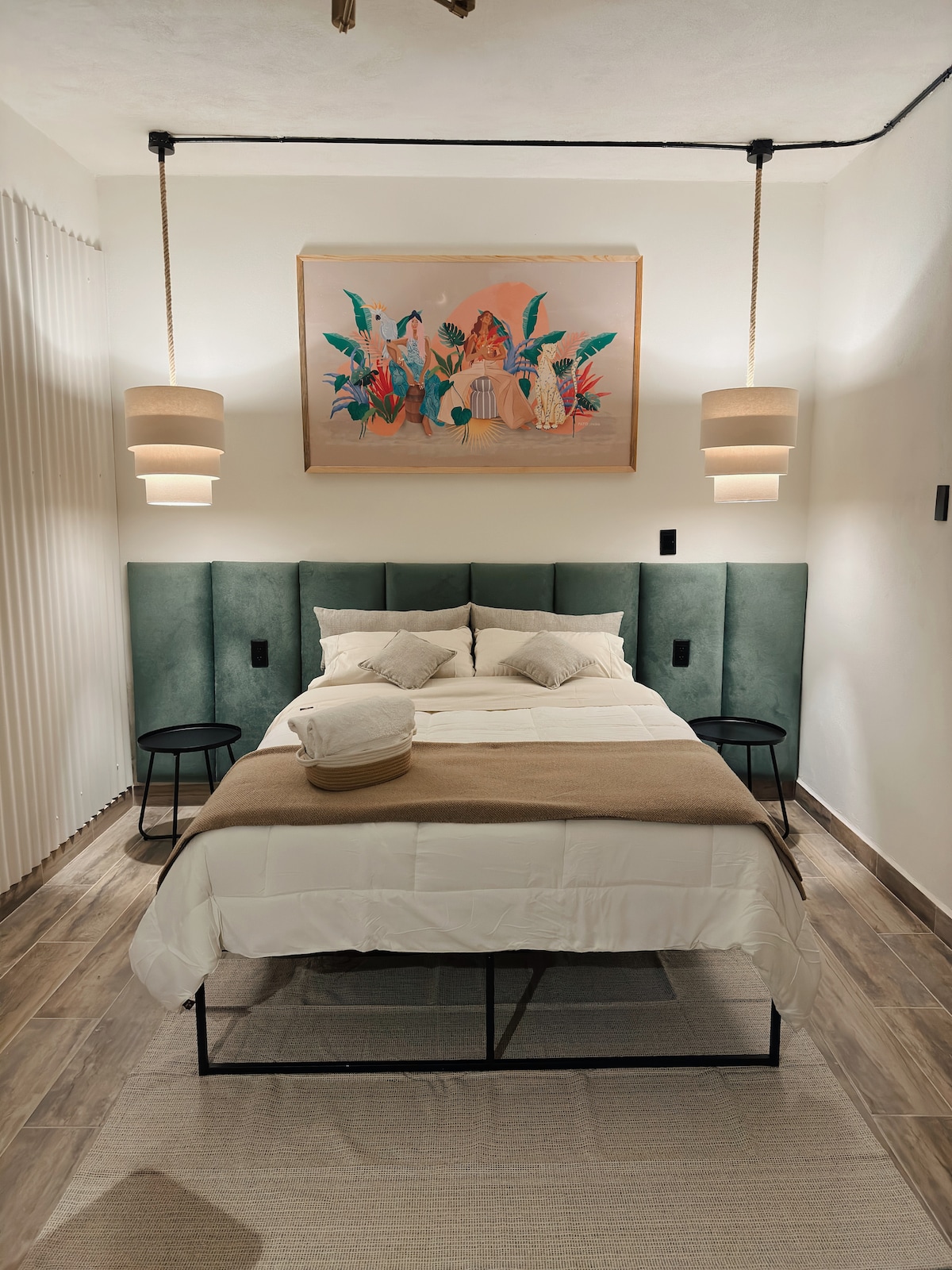 A cozy sleeping area features a spacious bed with soft linens and decorative pillows. Ambient light is provided by modern pendant lamps on either side. A colorful artwork adorns the wall above the bed, and two small side tables are positioned adjacent.