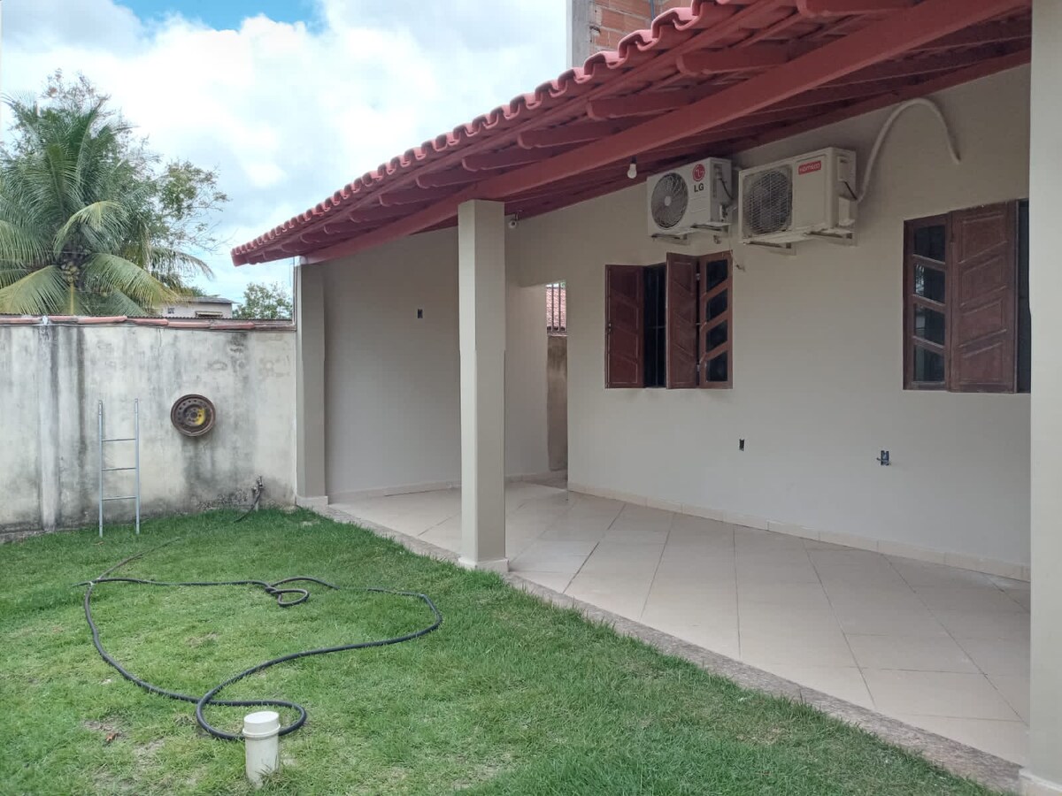 An outdoor area is presented with a well-maintained lawn and a covered patio. Air conditioning units are mounted on the wall, and a garden hose is coiled nearby. The space is bordered by a wall, offering a sense of privacy.