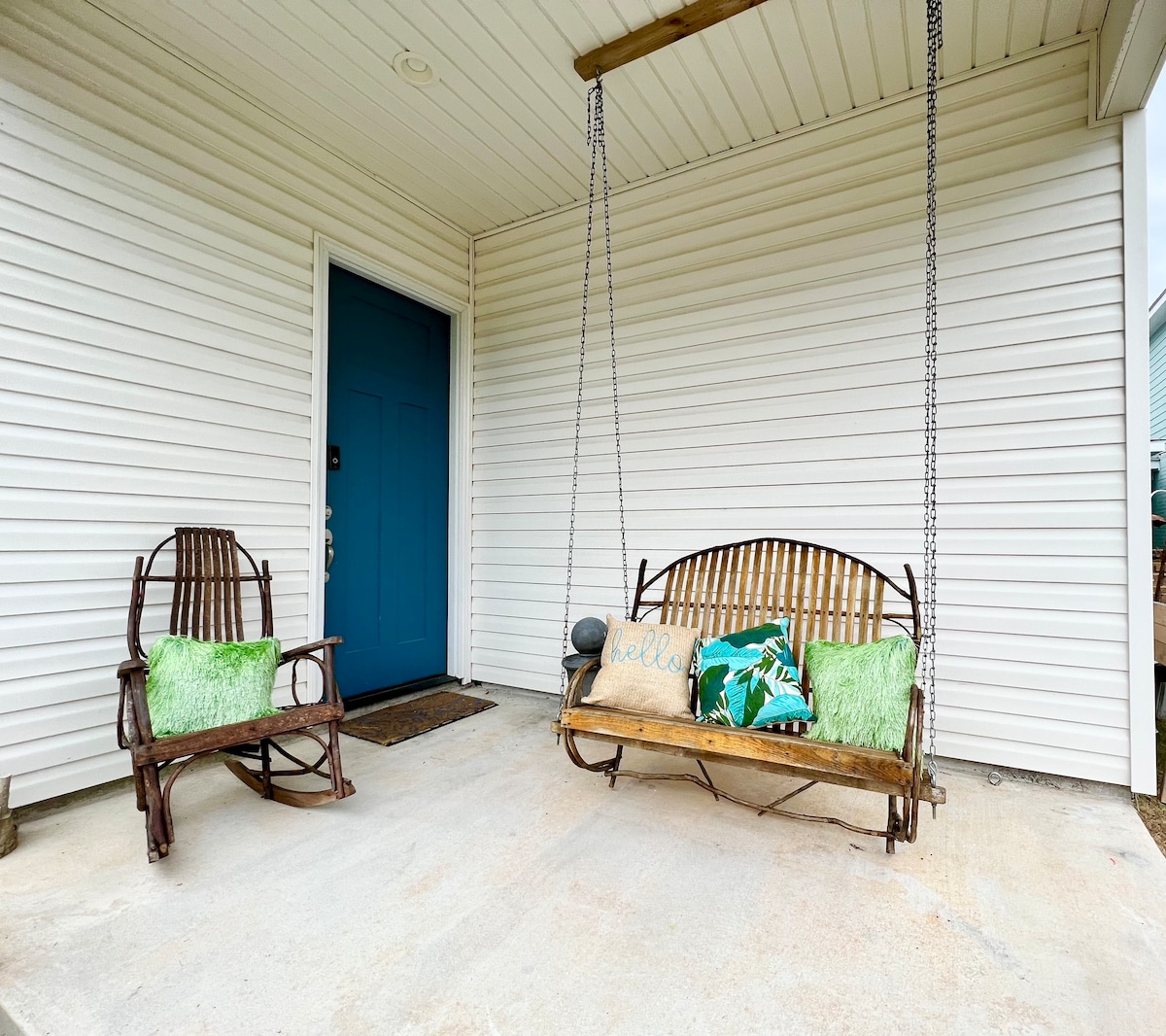 A welcoming porch features a rustic swing adorned with a decorative 'hello' pillow and two green accent pillows. A matching rocking chair sits nearby, adding charm to the entryway, which is framed by light-colored siding and a vibrant blue front door.
