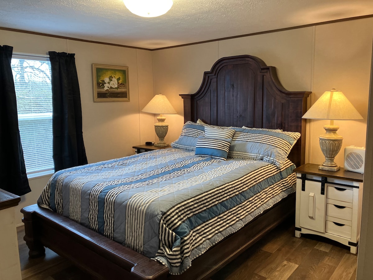 A spacious bedroom features a large wooden bed with a striped blue and white quilt and several pillows. Two bedside lamps provide soft lighting beside the bed. A window dressed with dark curtains allows natural light, and a piece of wall art adds a touch of decor.