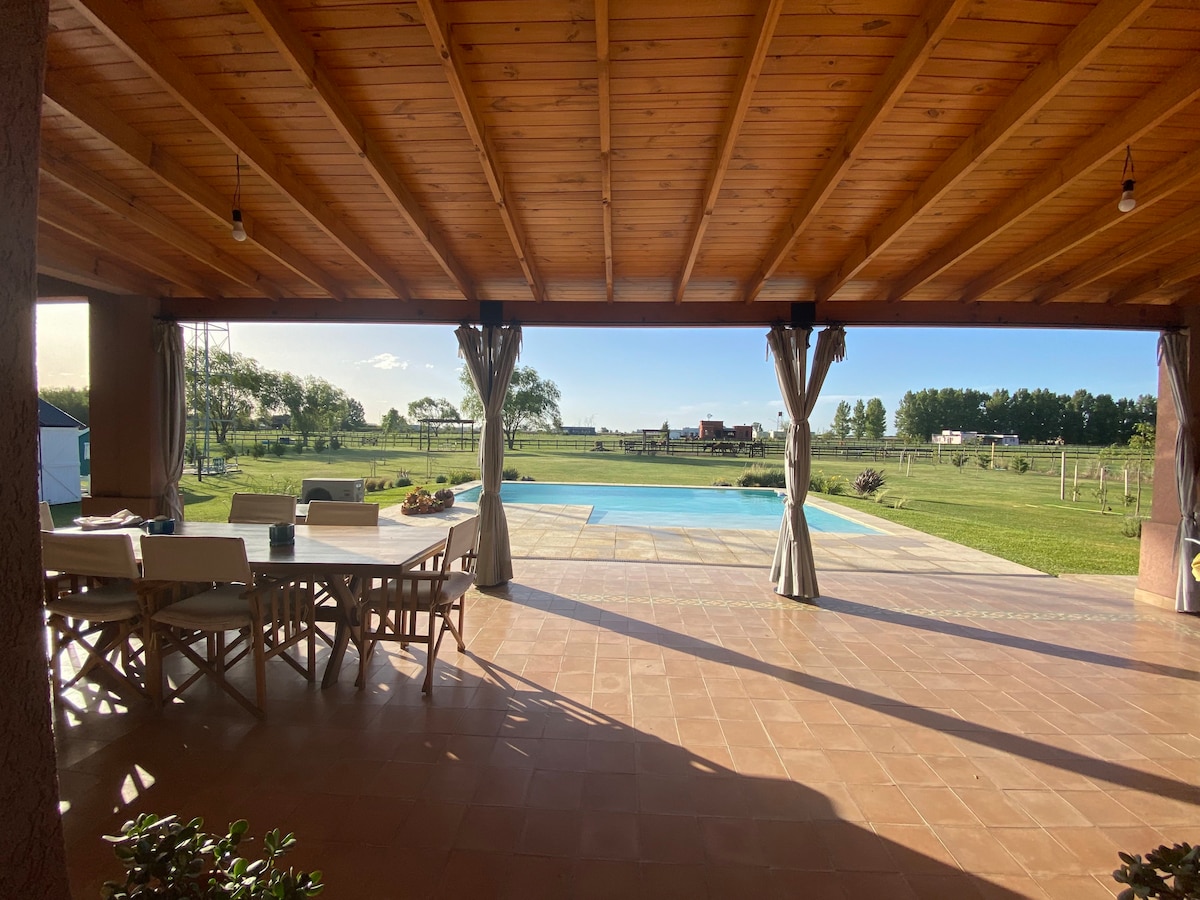 An outdoor covered patio features a spacious dining table surrounded by chairs, providing a view of the swimming pool and expansive green landscape. Two curtains frame the space, and sunlight illuminates the area, highlighting the natural surroundings.
