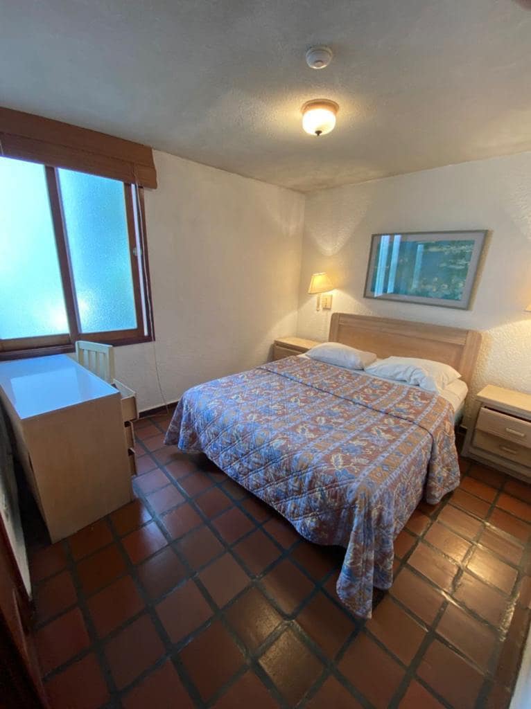 A well-lit bedroom features a spacious bed with a colorful patterned blanket. Two bedside tables with lamps are positioned on either side of the bed. A mirror is mounted above the bed, and a window allows natural light to fill the room.