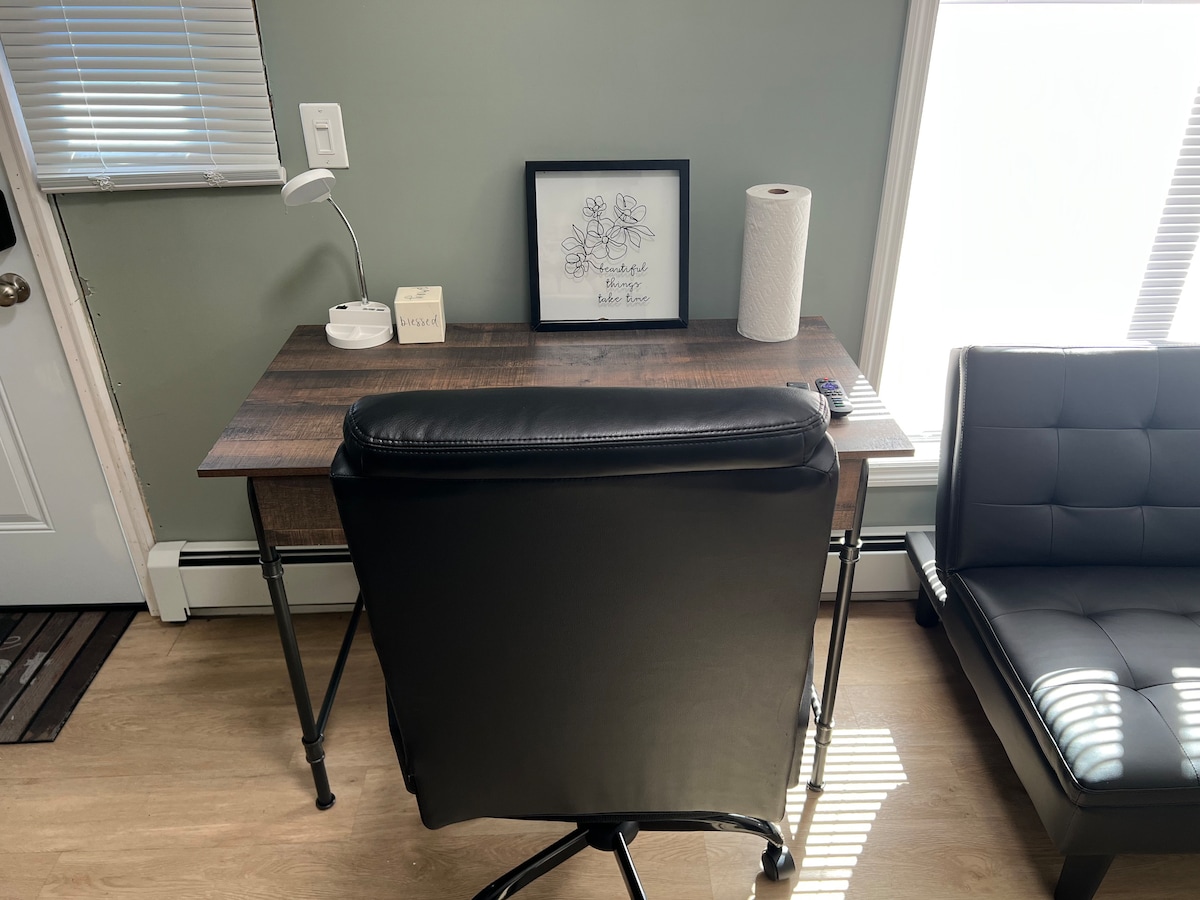 A functional workspace is showcased with a wooden desk and an ergonomic black chair. A table lamp provides illumination, while a framed artwork rests on the desk. A roll of paper towels and a light-colored box are also present, with natural light filtering through blinds.