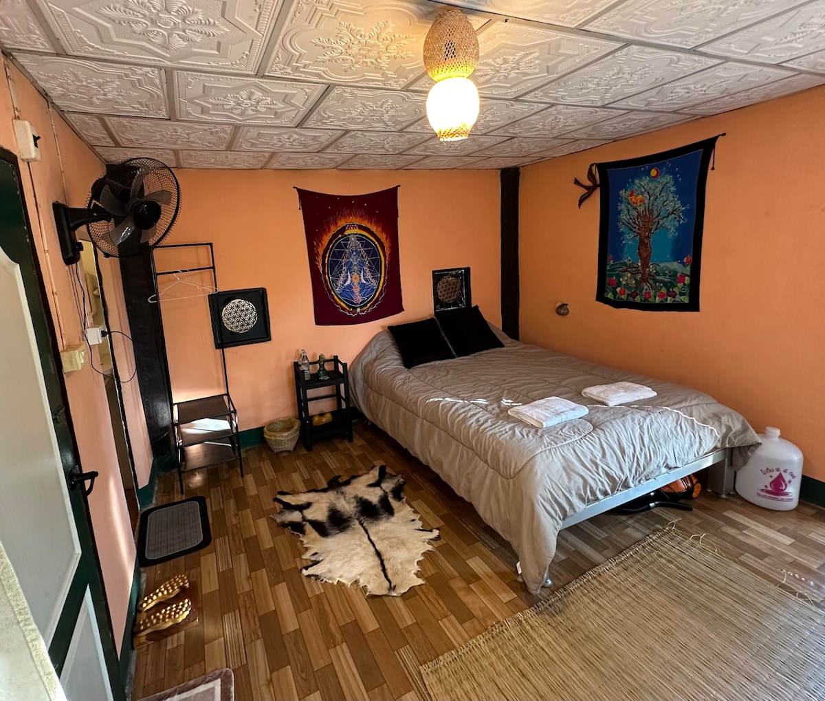 A serene sleeping area is highlighted by warm peach-colored walls and a textured ceiling. A spacious bed is dressed with soft linens and positioned against the wall. Two woven tapestries add color, while a natural rug lies on the wooden floor, complementing the overall rustic ambiance.