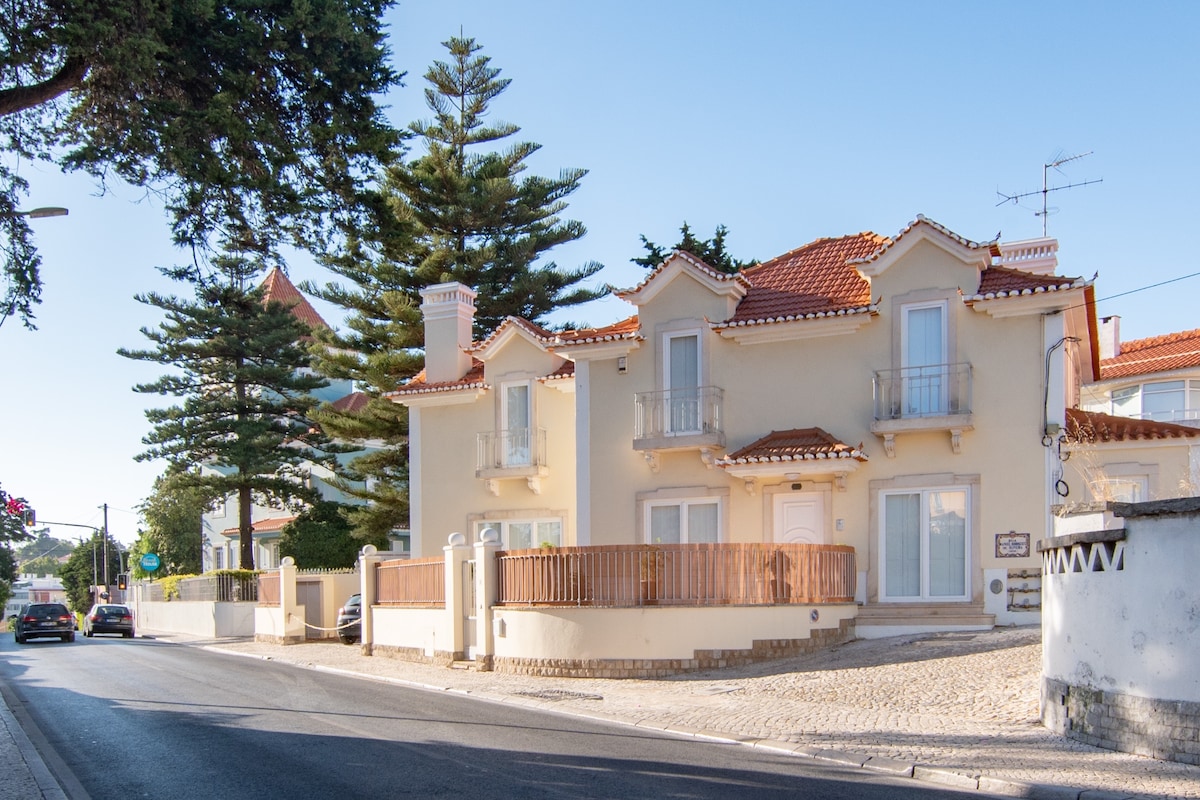 A charming multi-story villa is displayed with cream-colored walls and a red-tiled roof. Balconies are visible from the upper level, framed by green trees. A wooden fence encloses a small patio area, with a driveway leading to the street. Cars can be seen along the road.