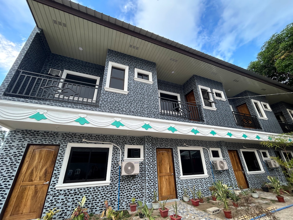 A two-story building is presented, showcasing a distinctive patterned facade with multiple windows. Air conditioning units are installed on both levels. Balconies with railings enhance the exterior, while potted plants add a touch of greenery along the stone path leading to the entrance.