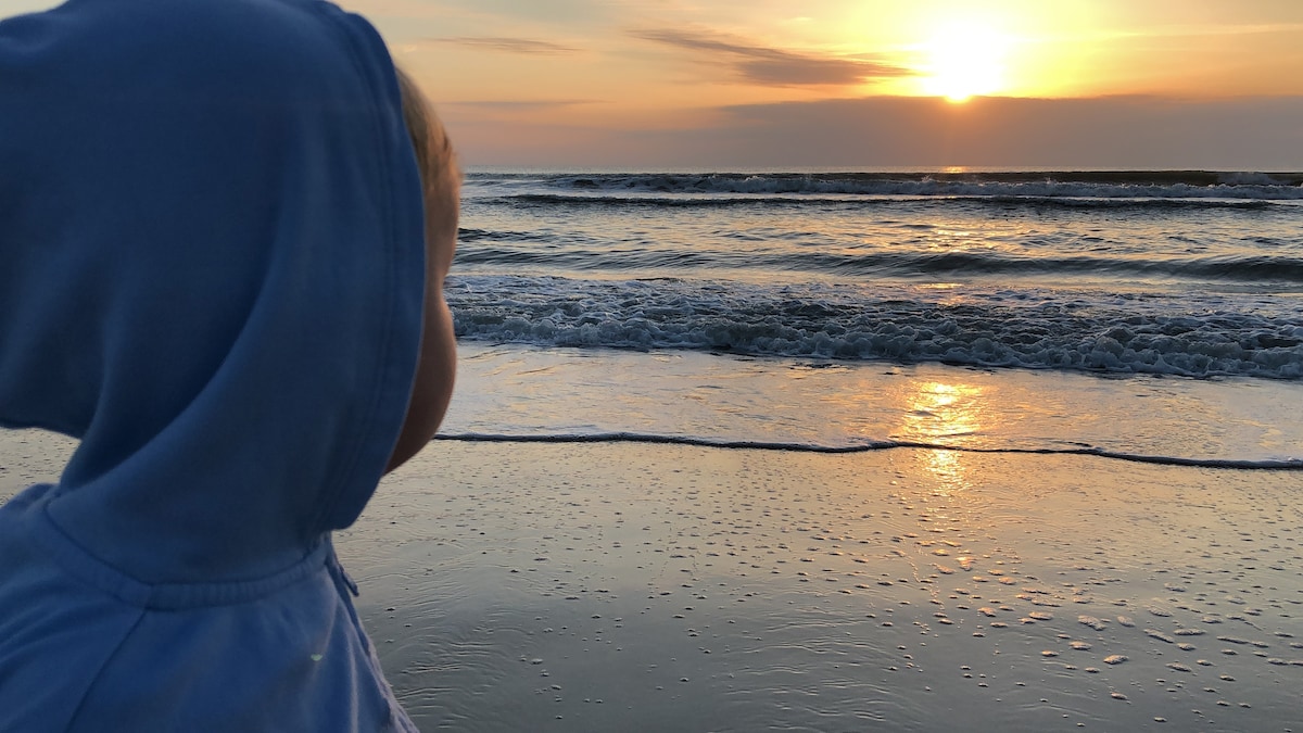 A child is observed gazing towards a sunset over the ocean, with gentle waves lapping at the shore. The water reflects warm hues of orange and gold, creating a serene backdrop against the sandy beach. The child's blue hoodie contrasts softly with the evening sky.