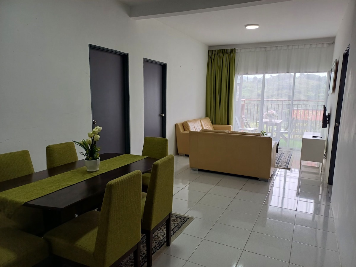 A bright living space features a light-colored sofa and a dining table with six green chairs. Large windows allow natural light to fill the room, with sheer curtains providing a light-filtering effect. A small balcony is visible through the sliding glass door.