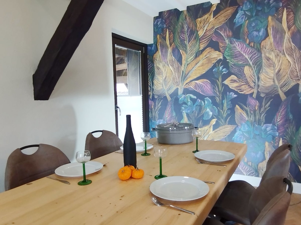 A dining area is presented with a wooden table accompanied by six chairs. The wall displays a vibrant floral mural, while a glass vase, bottle, and plates are arranged neatly on the table. Natural light enters through a nearby window, enhancing the space.