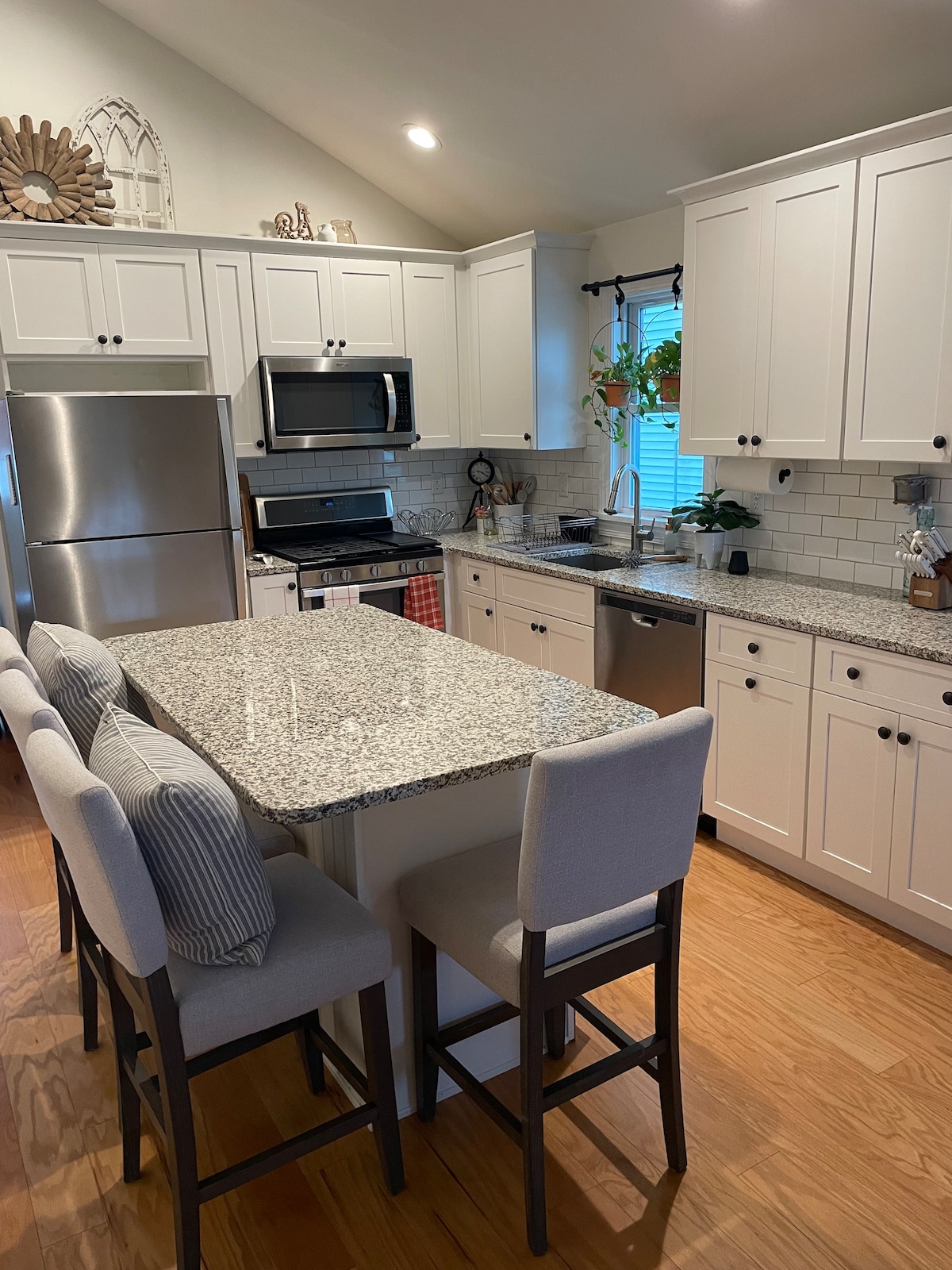 The kitchen features a large island with seating for four, complemented by granite countertops. Stainless steel appliances, including a refrigerator and microwave, are visible among white cabinets. Natural light filters through a window, illuminating the space and accenting plants on the countertop.