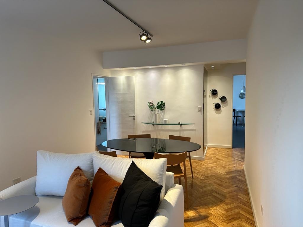 A light-filled living area is showcased with a white sofa adorned with dark cushions. An elegant round table with black chairs is positioned near a wall that highlights subtle, modern décor. The space features warm wood flooring, enhancing the inviting atmosphere.
