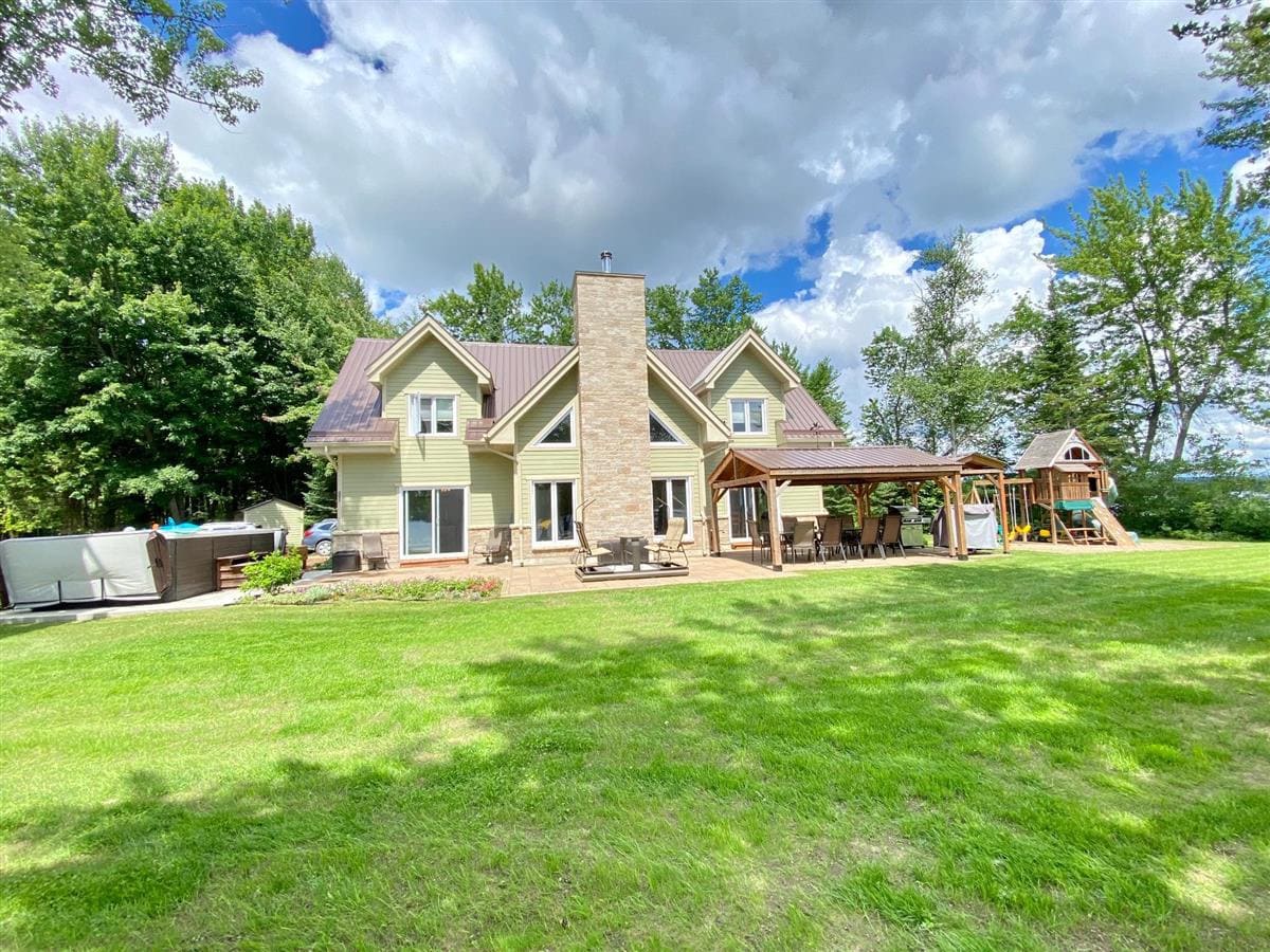 A spacious waterfront property features a large yard covered in lush green grass. The house, designed with a combination of stone and siding, has multiple gabled roofs. A shaded patio area with seating and a play structure are visible. Trees border the property, providing a natural setting.