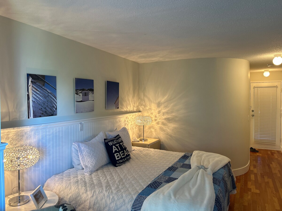 A bright sleeping area features a spacious bed dressed in white linens and accented with a blue throw. Flanking the bed are two unique nightstands with decorative lamps. Coastal-themed artwork adorns the wall above the bed, and natural light fills the room from the nearby entrance.