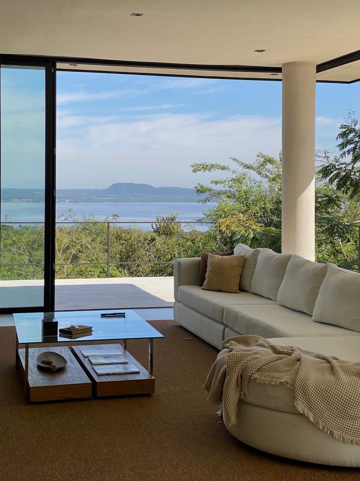 A spacious living area is presented with large glass doors, offering a view of the lake and surrounding greenery. A light-colored sectional sofa is complemented by a soft blanket draped over it. A glass coffee table sits in front, with a few magazines and a remote on its surface.