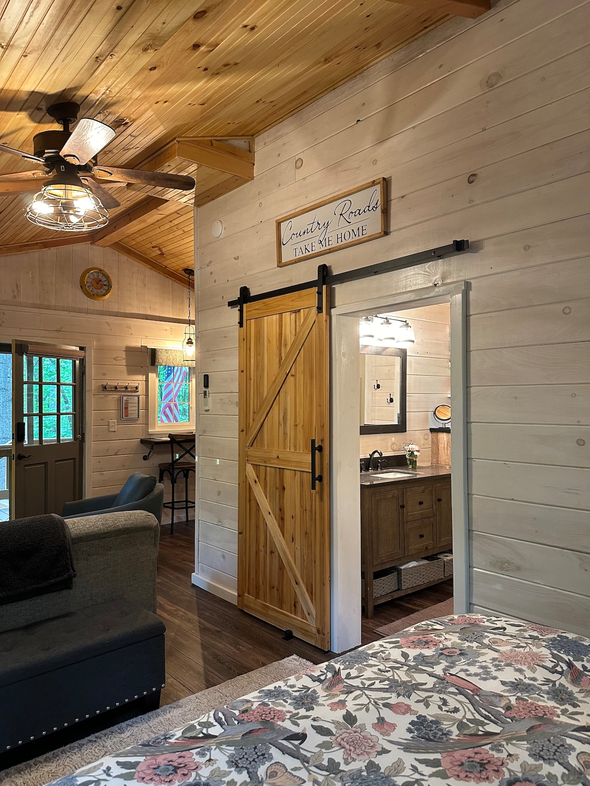A cozy cabin interior features wooden walls and ceiling accents. A sliding barn door leads to a bathroom space, and a comfortable couch is positioned nearby. Artwork and decorative elements add character, while a ceiling fan is visible, enhancing the inviting ambiance of the room.