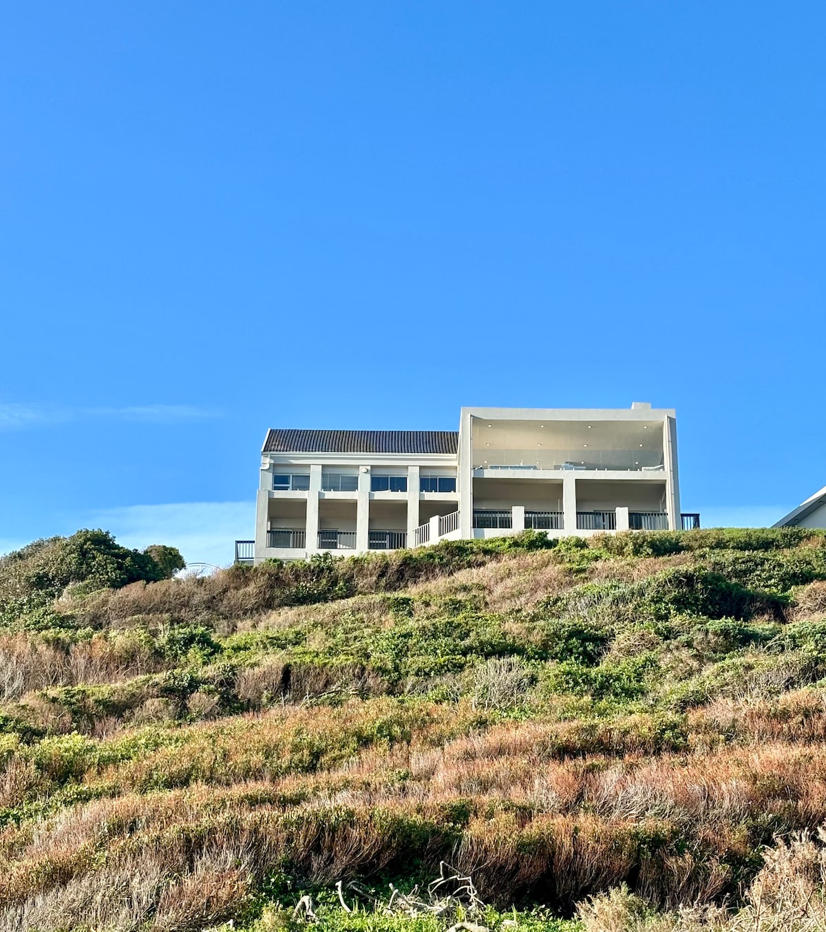 A spacious beach house is seen from below, set on a ridge with several floors visible. A clear blue sky serves as the backdrop, while the lush greenery of the slope surrounds the property. Large balconies can be observed, enhancing the home's outdoor space.