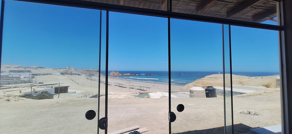 A large glass window offers an unobstructed view of the coastline, revealing sandy beaches and rolling waves. The landscape features gentle hills in the background, with a clear blue sky above, creating a serene beach atmosphere.