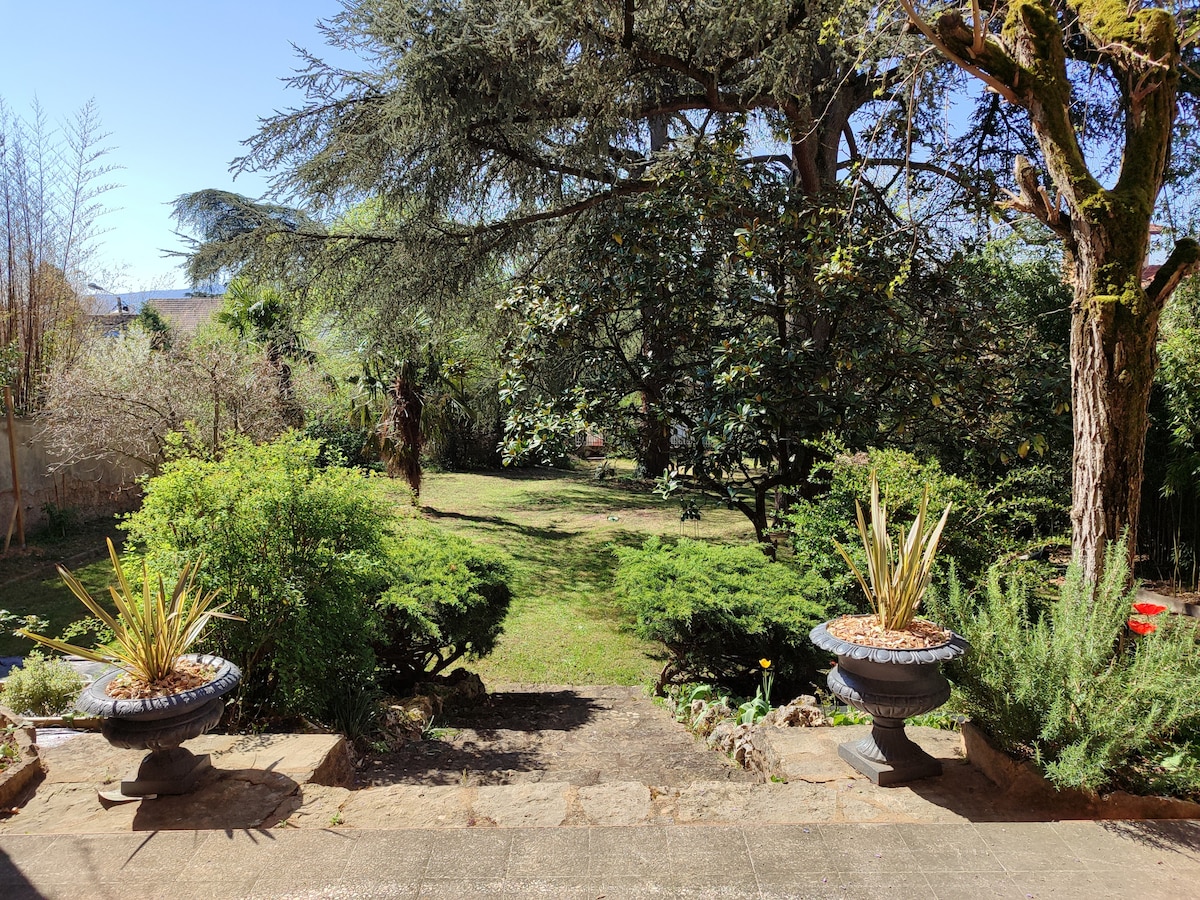 A spacious garden is visible, featuring lush green grass and a variety of well-maintained plants and trees. Stone steps lead down from a terrace, framed by ornamental planters. The serene outdoor setting is surrounded by foliage, providing a peaceful atmosphere.