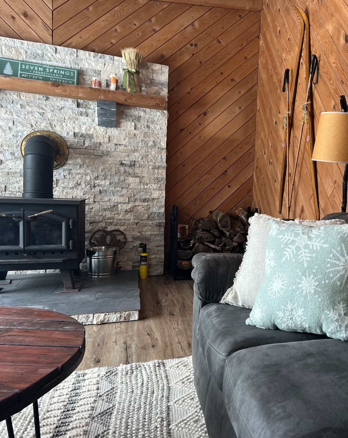 A stone fireplace serves as a focal point in a cozy living area, complemented by wood paneling. A comfortable gray sofa adorned with a light teal pillow sits in front of the fireplace, with a wooden coffee table positioned nearby. A stack of firewood is neatly arranged beside the hearth.