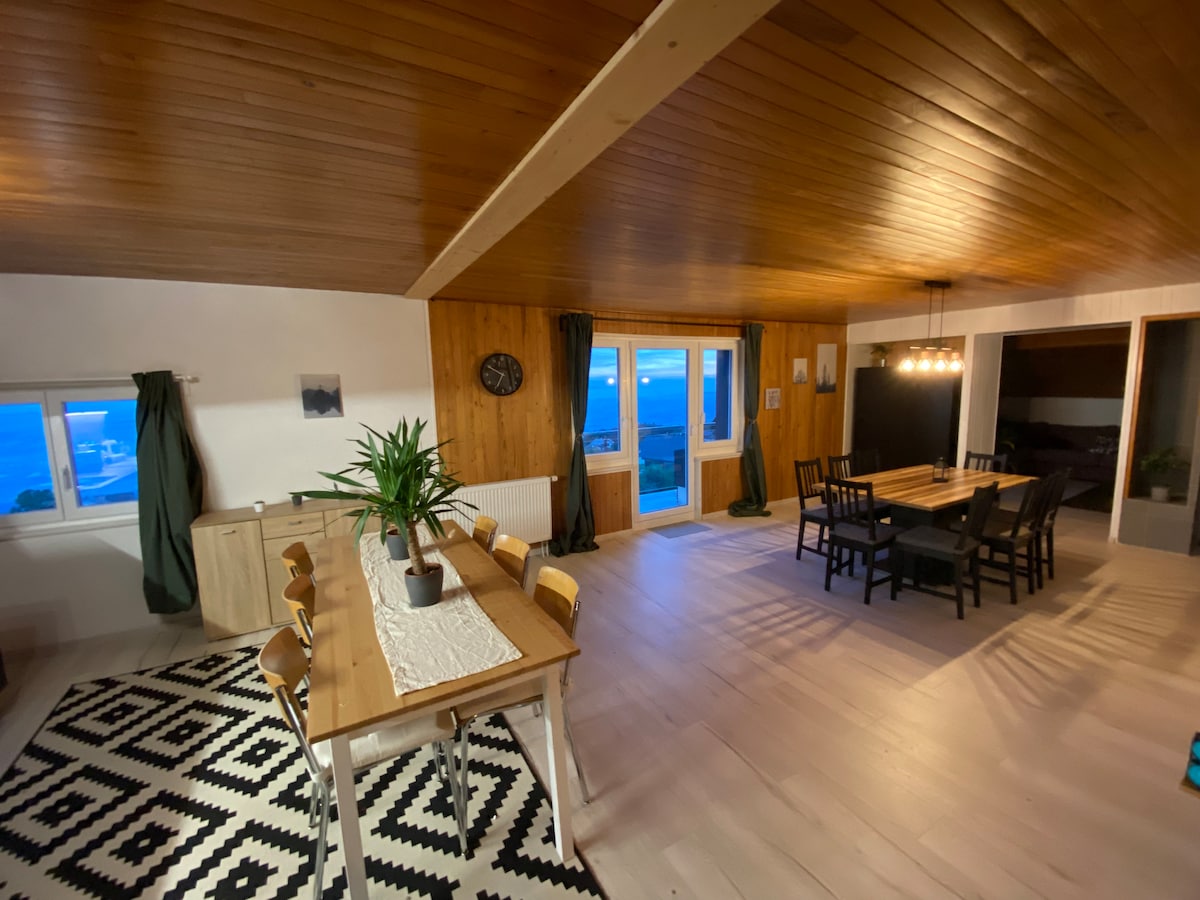 A spacious dining area features a long wooden table surrounded by dark chairs, illuminated by pendant lights. Large windows offer a view of the outside, while a patterned rug adds warmth to the light-colored floor. A clock is mounted on the wall, enhancing the decor.