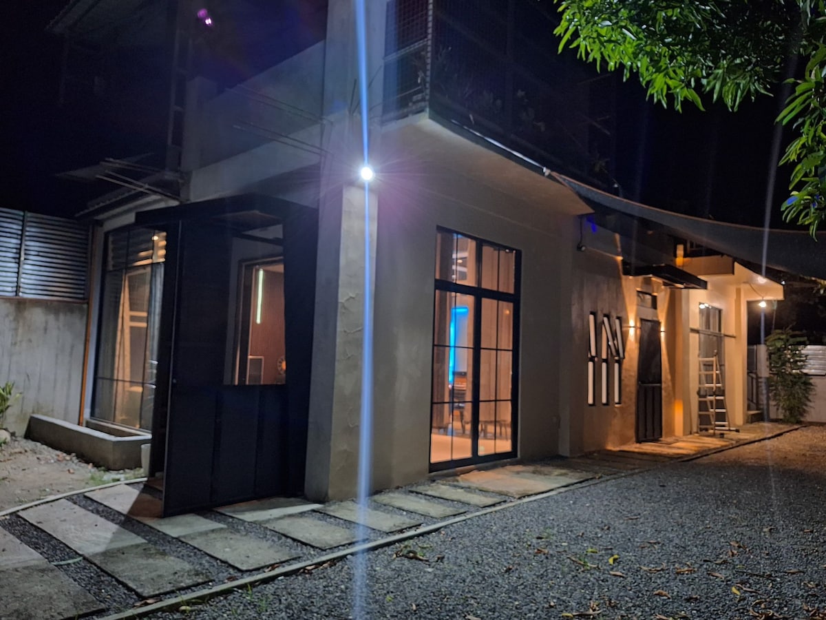 The exterior of the residence is highlighted at night, featuring large windows that reflect soft interior lighting. A gravel pathway leads to the entrance, flanked by structured landscaping and illuminated by subtle outdoor lighting.