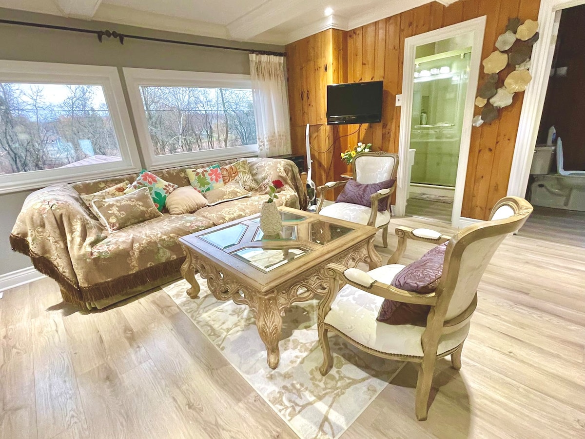 A cozy living area is highlighted by a comfortable sofa adorned with colorful cushions. A glass coffee table sits centrally, surrounded by elegant armchairs. Natural light pours in through large windows, while warm wood paneling adds a touch of rustic charm to the space.