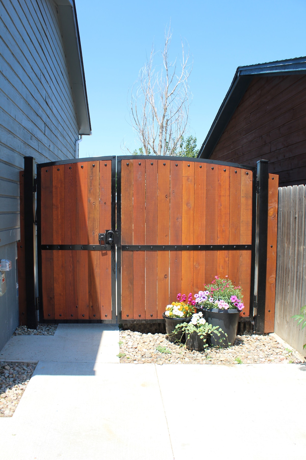 A sturdy wooden gate made of horizontal planks is framed in black metal. It is positioned between two buildings, leading to a narrow pathway. Colorful potted flowers add a touch of greenery at the base of the gate, enhancing the inviting entrance.