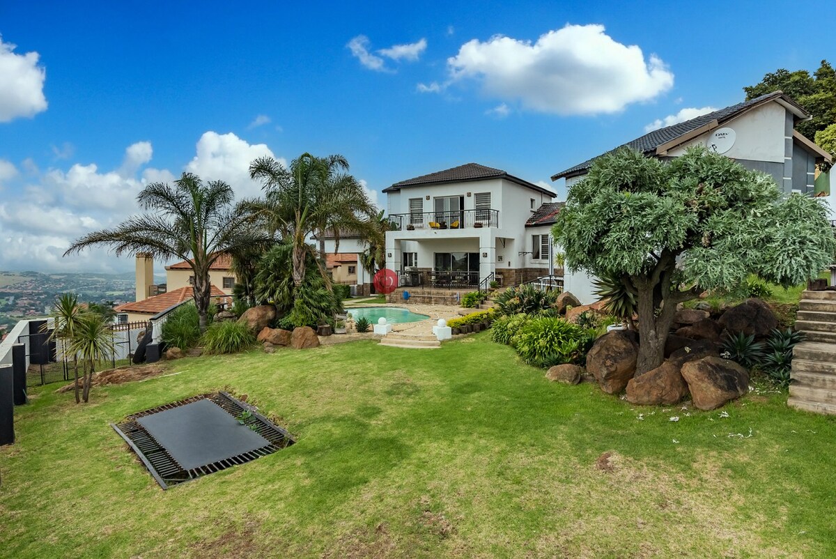 A spacious backyard features lush green grass and ornamental plants surrounding a swimming pool. A patio area is visible on the house, while stone pathways and steps lead to various levels of landscaping, enhancing the natural setting.