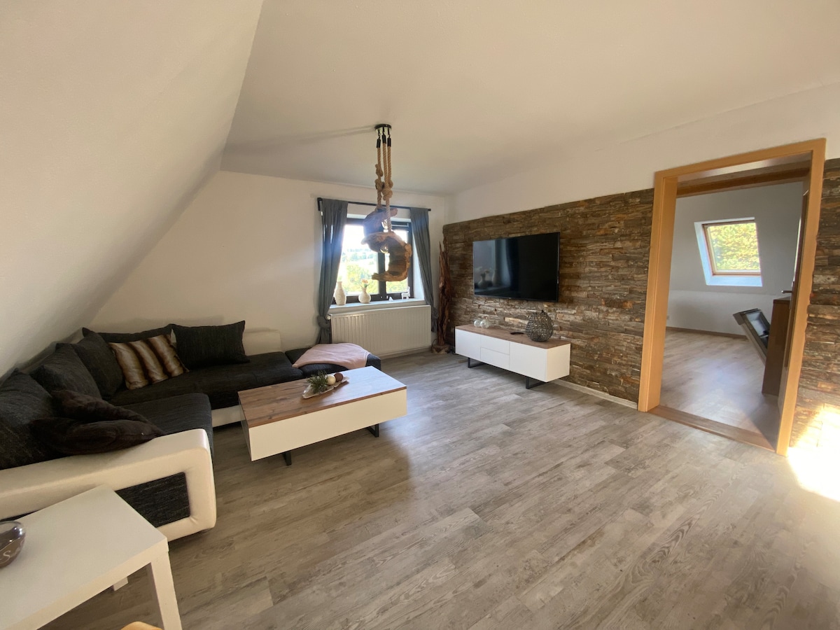 A spacious living area is seen, featuring a sectional couch with cushions and a coffee table. A wall-mounted television is positioned across from the seating. Natural light enters through a window, highlighting a textured stone accent wall and a doorway leading to another room.