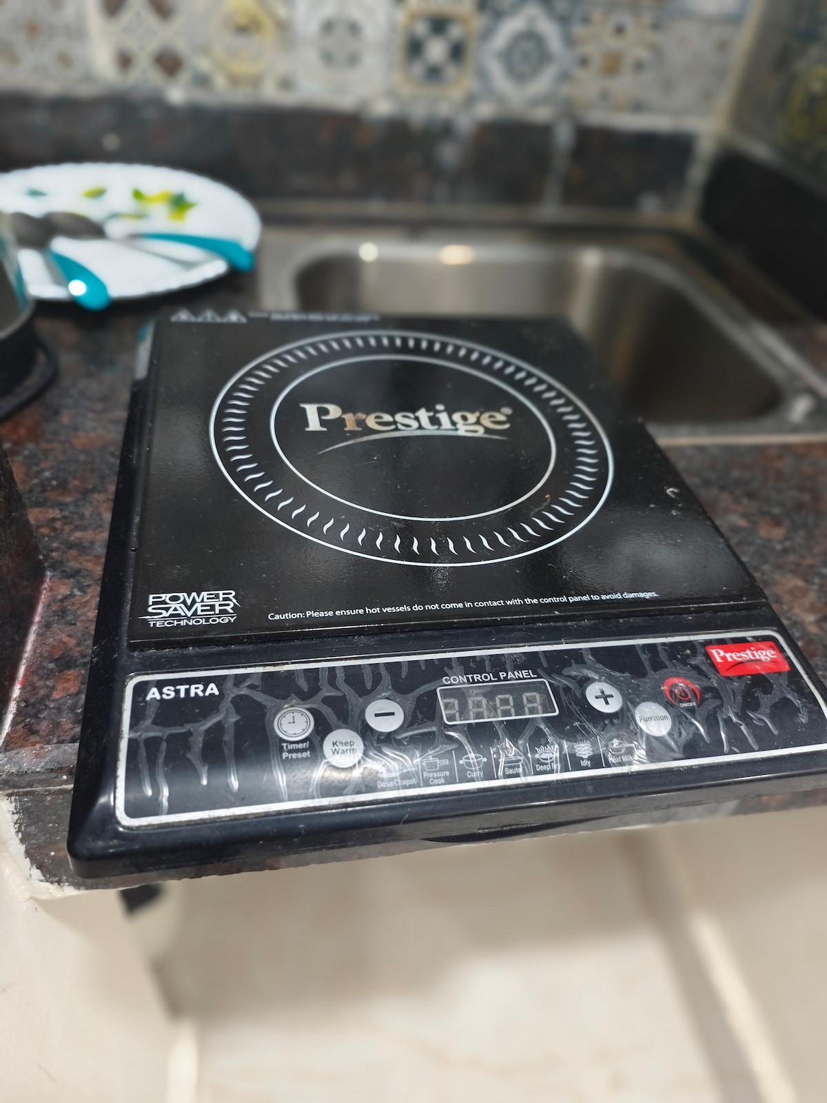 A portable induction cooktop is displayed on a kitchen countertop, featuring a sleek design with a control panel and digital display. The countertop, made of dark stone, includes a stainless steel sink in the background, suggesting a functional kitchen space.