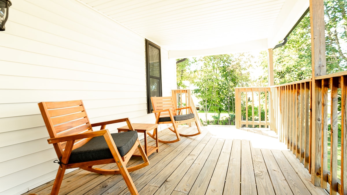 A welcoming outdoor deck area is depicted, featuring two wooden rocking chairs positioned on a wooden floor. Sunlight filters through the surrounding trees, illuminating the space. The gentle curvature of the deck enhances the sense of openness and connection to nature.