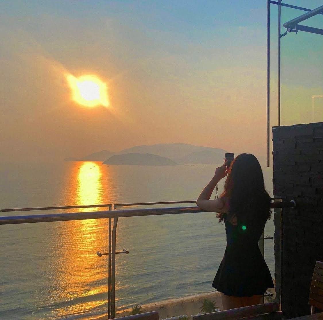 A stunning view is visible from the balcony, featuring a golden sunset reflecting on the calm sea. The silhouette of a person stands captured while taking a photo, framed by the balcony railing and the vast landscape beyond.