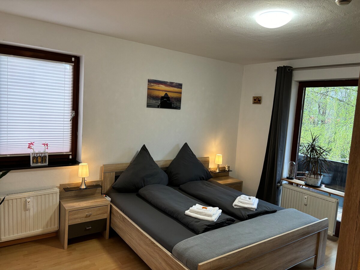 A spacious bedroom is presented with a large bed featuring two oversized pillows. Nightstands are positioned on either side, each with a soft-lit lamp. A landscape photograph adorns the wall above the bed. Natural light enters through a window dressed with light blinds, revealing a glimpse of greenery outside.
