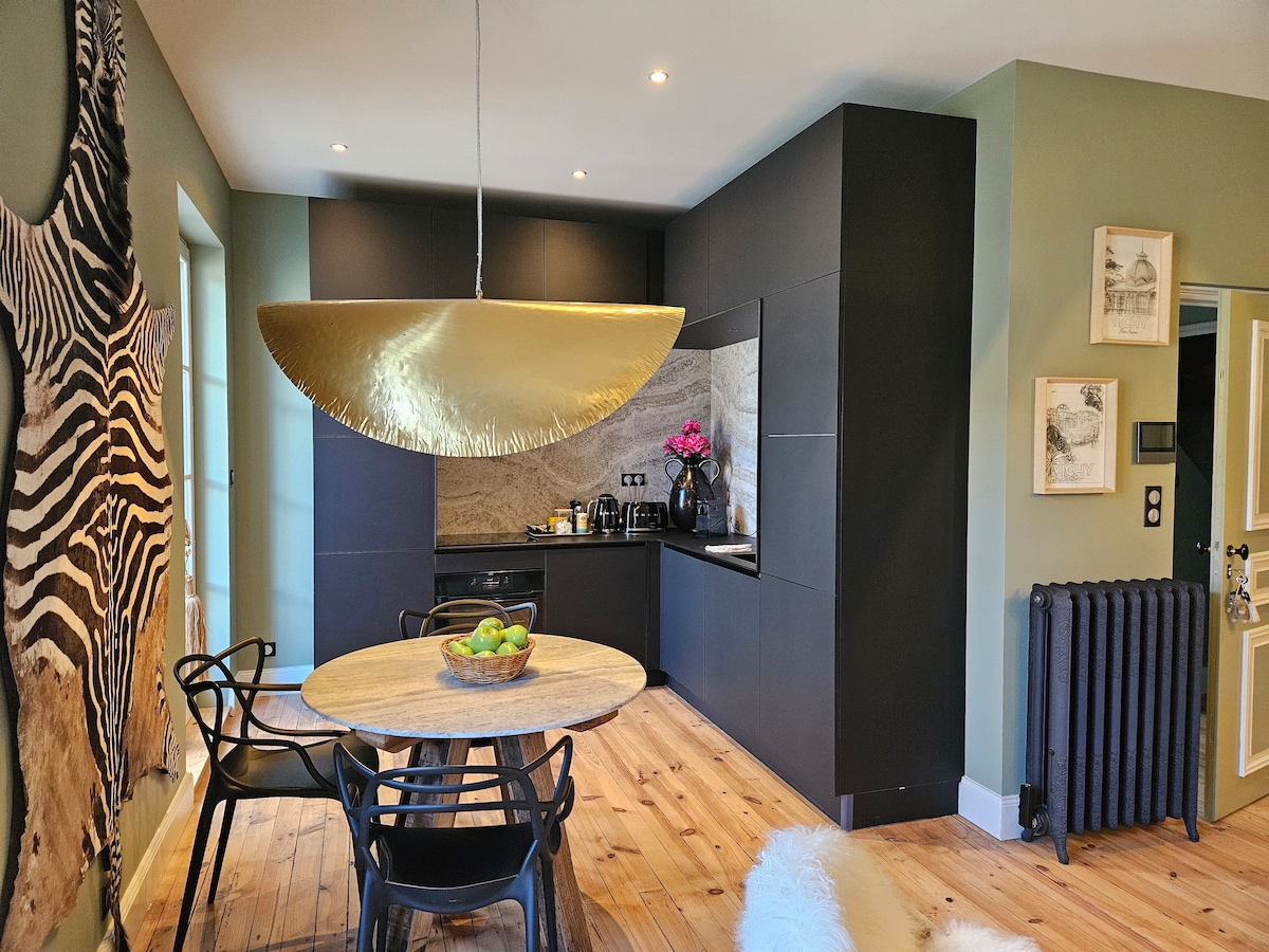 A modern kitchen area features a sleek black cabinetry with a stylish gold pendant light overhead. A round wooden table is surrounded by four black chairs, while fresh fruit is displayed in a bowl at the center. A textured wall art piece adds character to the space.