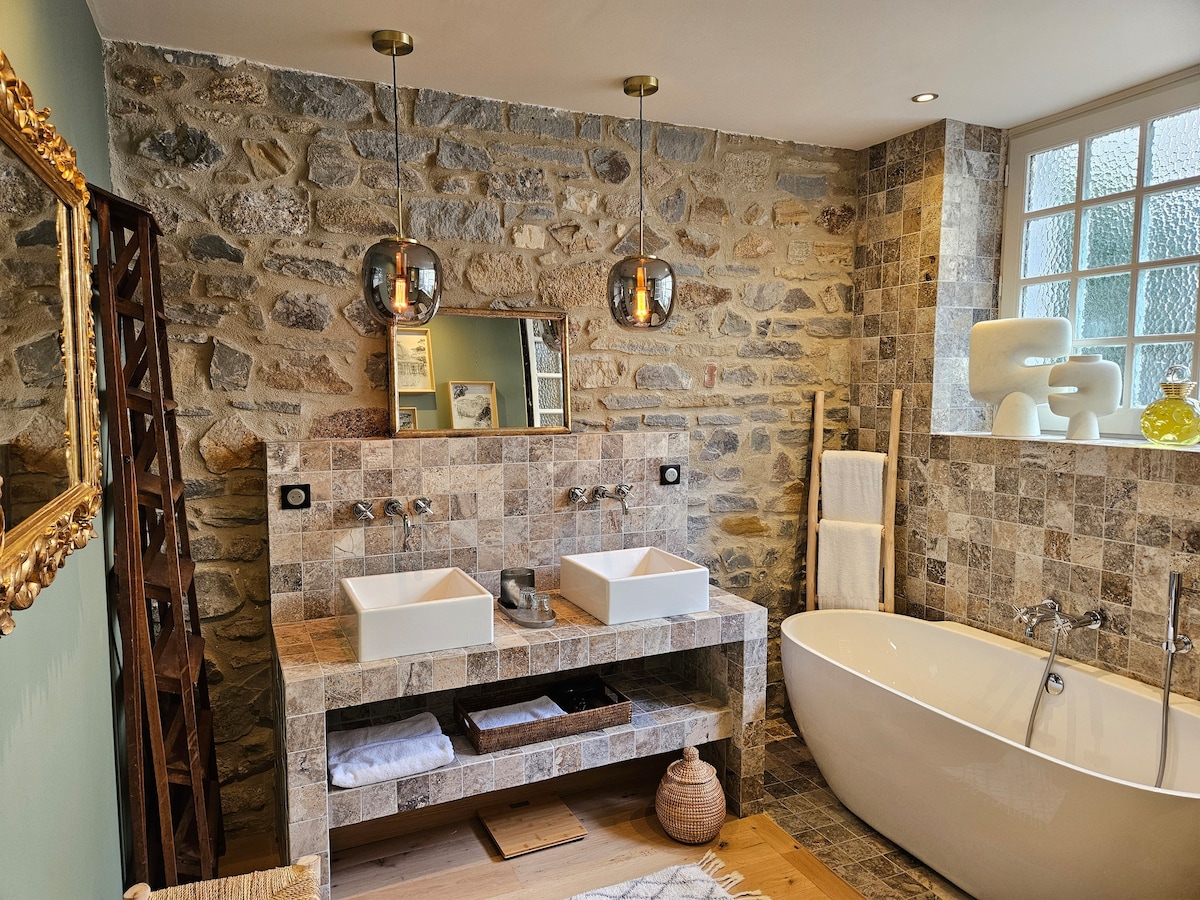 A spacious bathroom features textured stone walls and elegant fixtures. Two square basins are placed atop a wooden countertop, complemented by modern pendant lights. A freestanding bathtub offers a relaxing focal point, while soft towels and decorative elements enhance the tranquil setting.