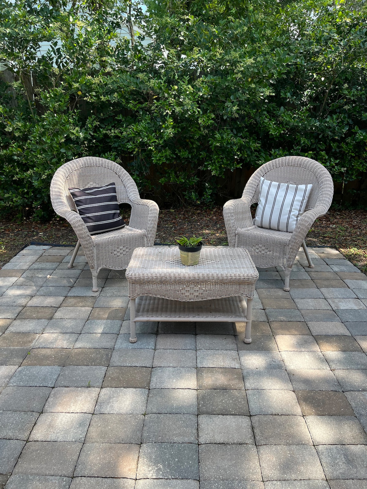 A cozy outdoor seating area features two woven chairs with accent pillows, paired with a coffee table. The space is paved with square stones, surrounded by lush greenery, creating a sense of privacy and tranquility.