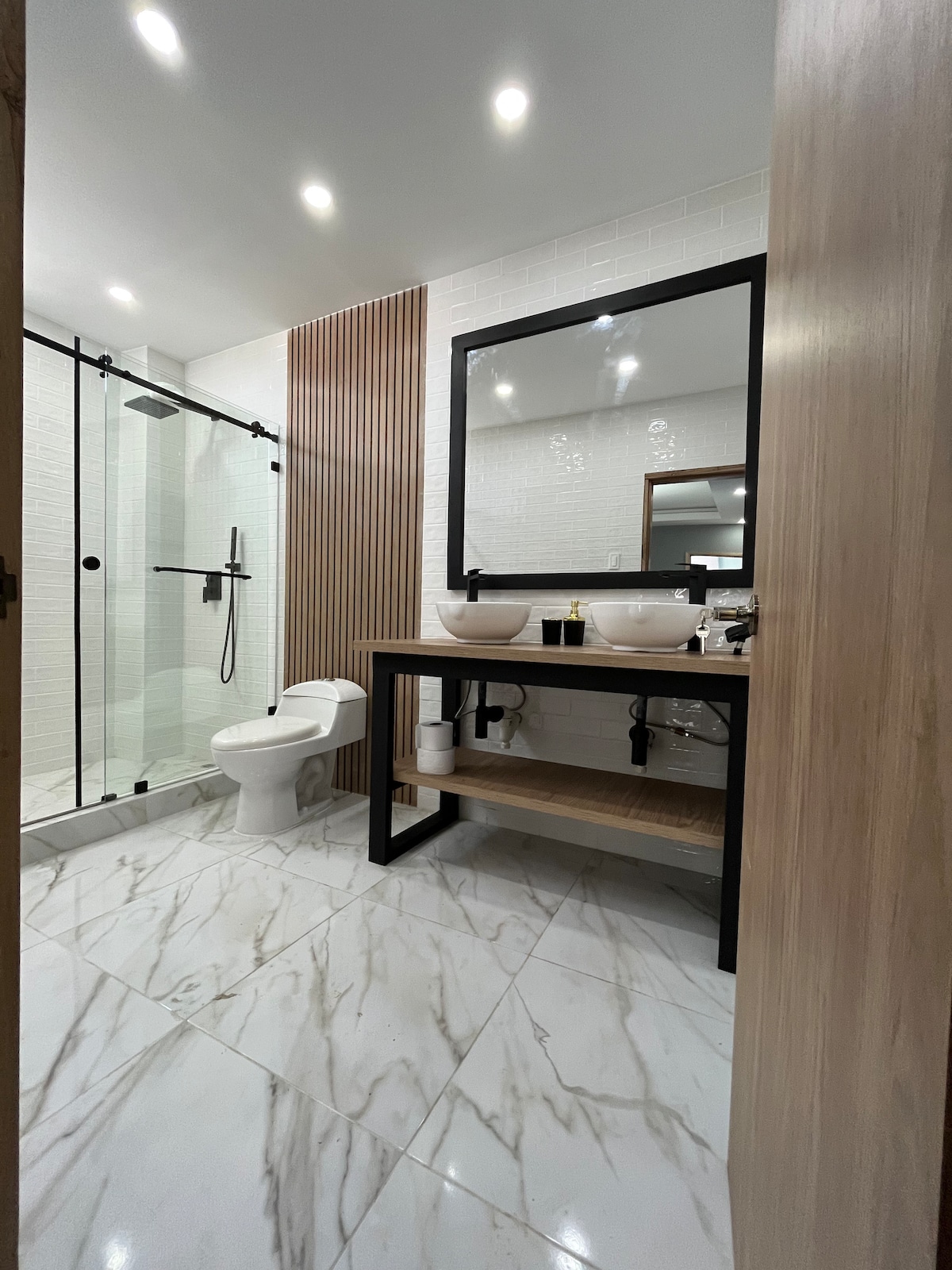An elegant bathroom features a walk-in shower enclosed by glass panels. Dual vessel sinks are positioned above a wooden vanity with open shelving. The floor and walls are adorned with marble tiles, while warm lighting enhances the modern design.