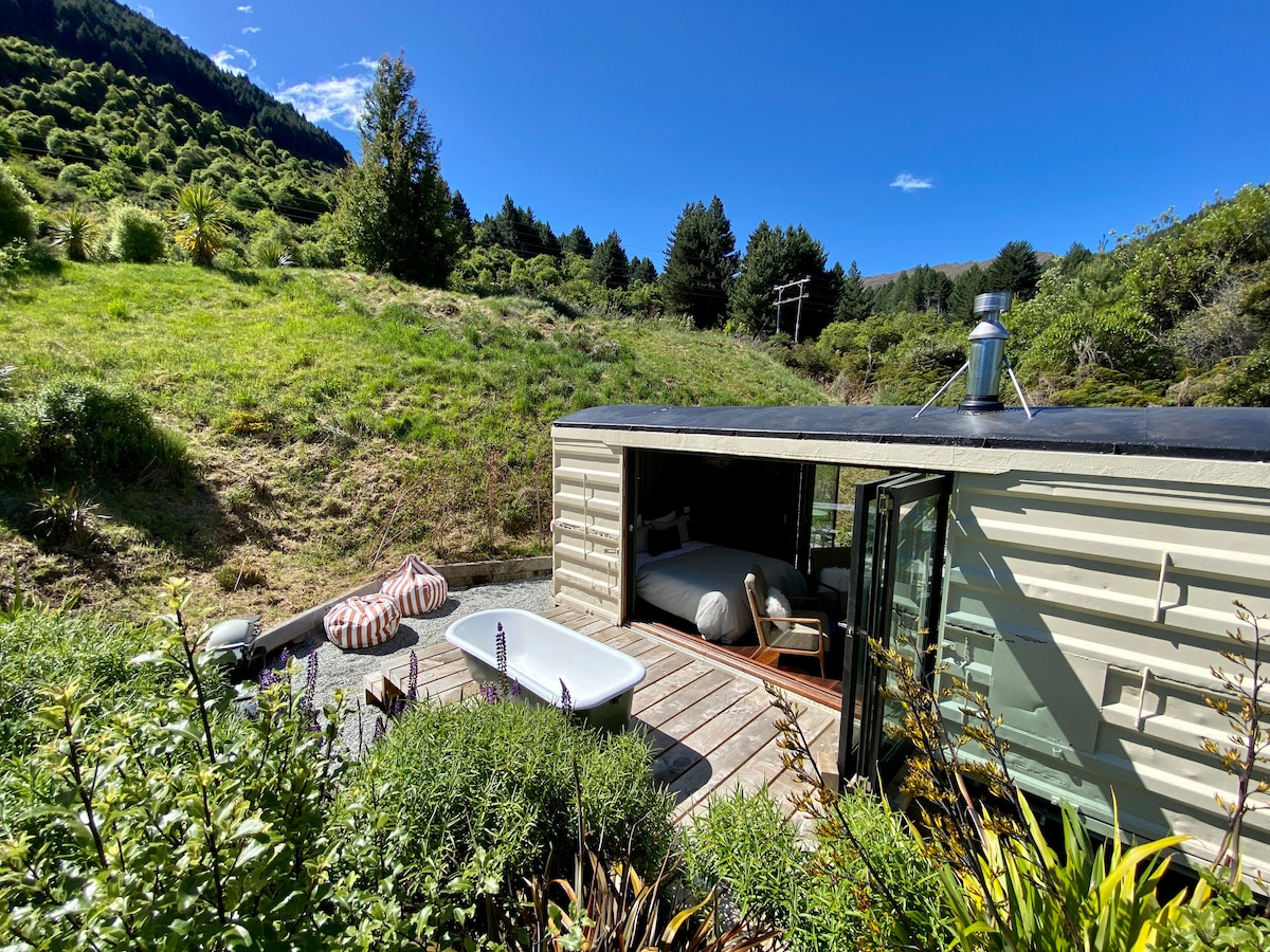 A retreat space is set amidst lush greenery, featuring a private outdoor bath on a wooden deck. A spacious bed is visible inside through large glass doors, while comfortable seating is positioned nearby. Natural light highlights the tranquil surroundings and scenic landscape.