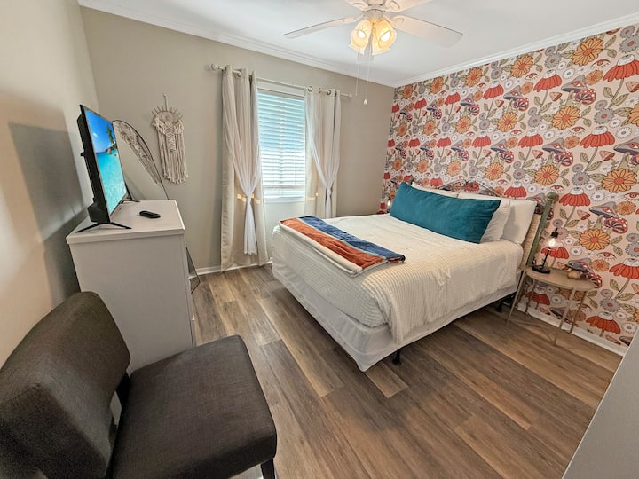 Bright bedroom with bold floral accent wall, Roku TV, ceiling fan and cozy queen bed. Dresser and closet for clothing storage.