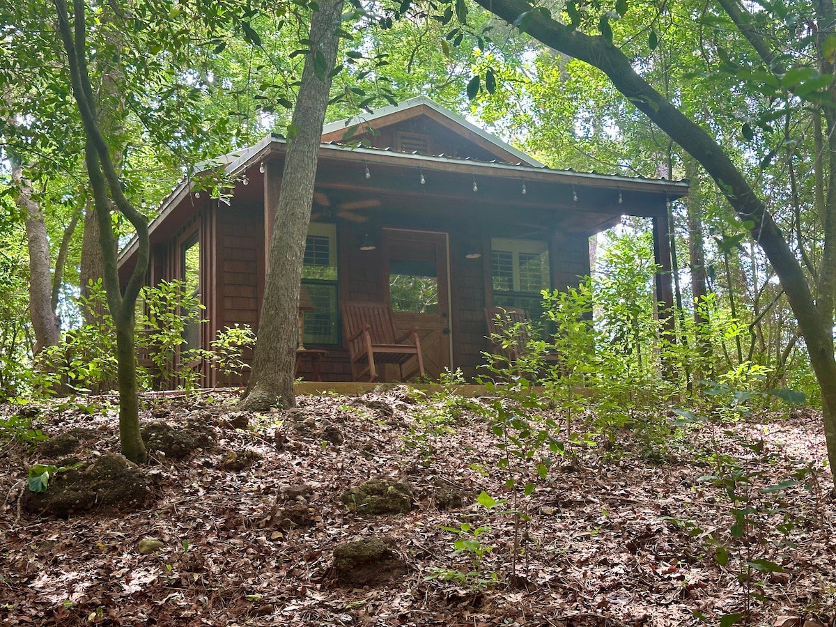 A rustic cottage is nestled among the trees, surrounded by a natural landscape of leafy foliage and earthy tones. A covered porch features two rocking chairs, offering a quiet space to relax. Sunlight filters through the branches, creating a serene atmosphere.