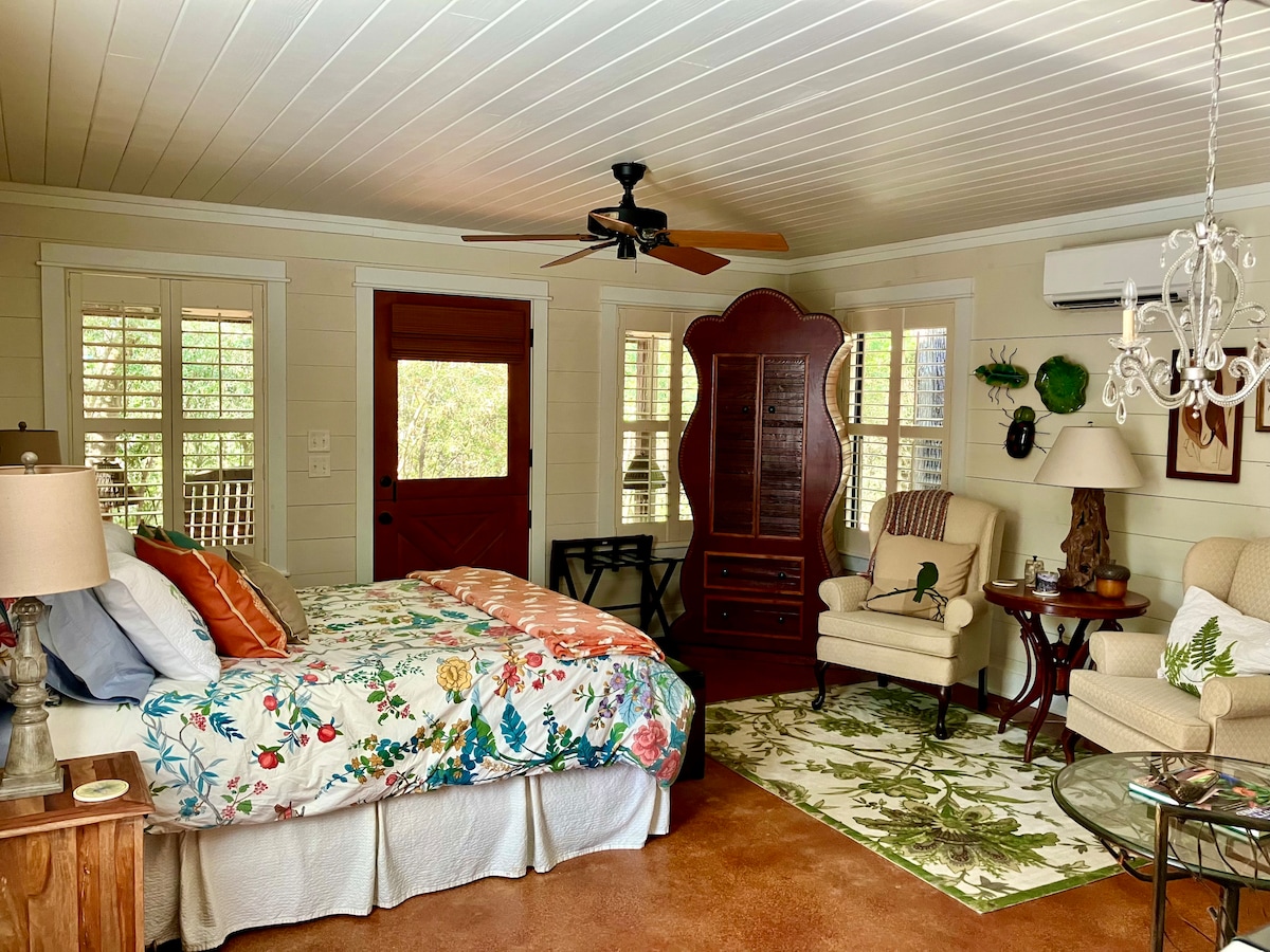 A cozy sleeping area is featured with a floral-patterned bedding set atop a spacious bed. The room includes a comfortable chair, a small side table, and a vintage dresser. Natural light filters through multiple windows, creating a serene atmosphere.