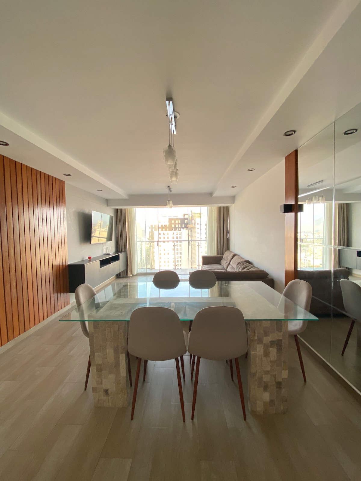 A modern dining area is depicted, featuring a glass table surrounded by four light-colored chairs. Natural light fills the space through large windows, accentuating the sleek design of the adjacent living room, which includes a gray sofa and wood paneling.