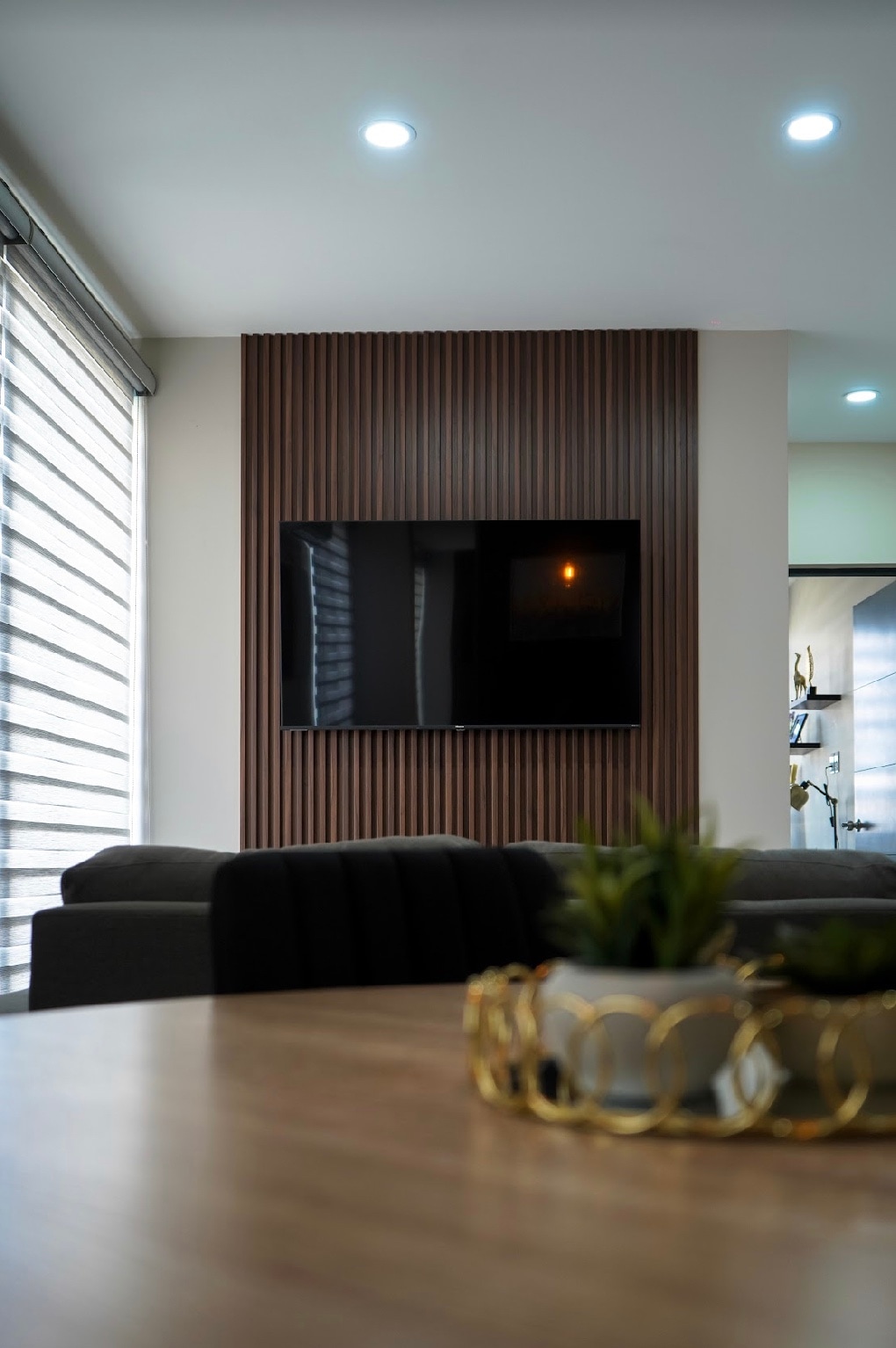 A modern living area features a sleek television mounted on a wooden accent wall. Soft lighting enhances the space, while a coffee table with a decorative plant provides a touch of greenery. Large windows are dressed with stylish blinds, allowing natural light to filter through.