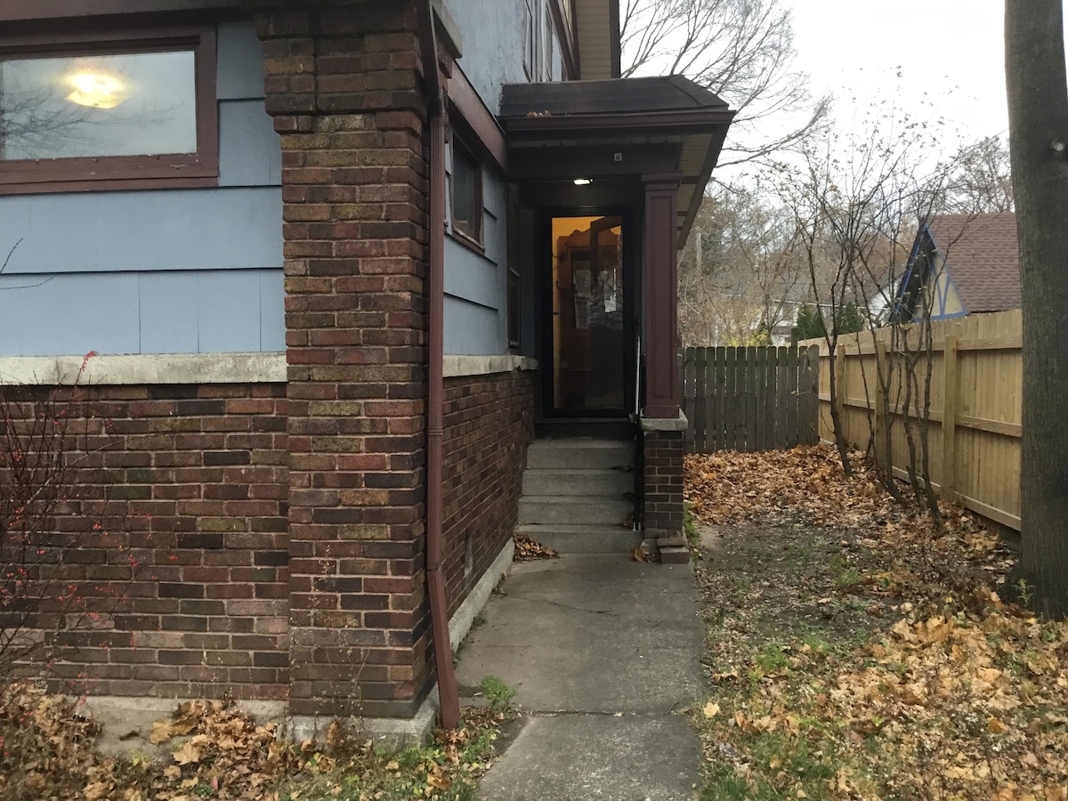 An exterior view of the house showcases a brick façade and blue siding. A concrete path leads to the entrance, which is framed by a small overhang. A private backyard is visible beyond a wooden fence, with fallen leaves scattered on the ground.