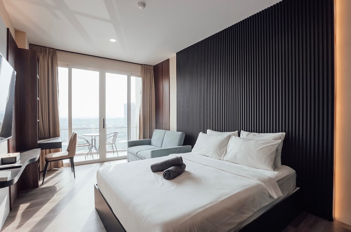 A modern bedroom features a spacious bed with white linens, complemented by a soft gray couch. Large windows provide natural light and access to a private balcony. A sleek desk and chair are positioned against the wall, enhancing the room's contemporary design.