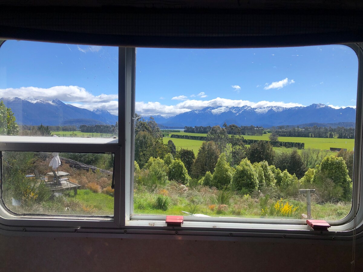 A panoramic view of lush green fields and the majestic Fiordland mountains is captured through the large window. Snow-capped peaks rise against a bright blue sky, enhancing the natural beauty of the serene landscape outside.
