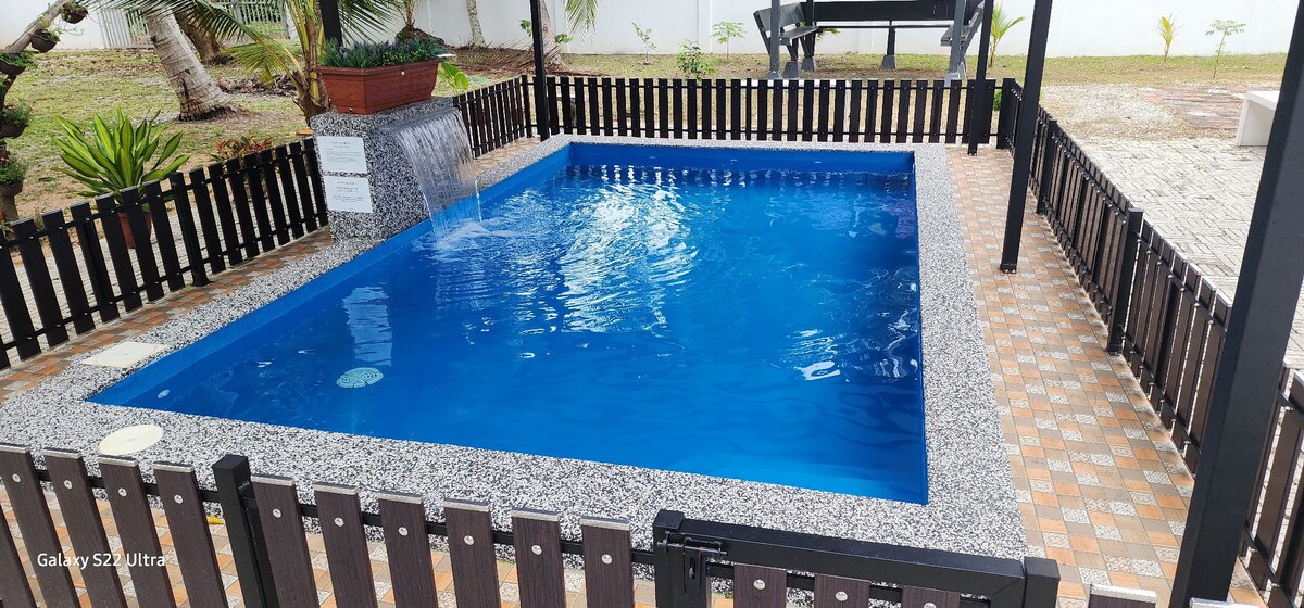 A serene outdoor space features a private pool surrounded by a low fence. The pool's clear blue water is complemented by a stone deck, while a gentle waterfall adds a calming sound. Lush greenery is visible in the background, enhancing the peaceful environment.