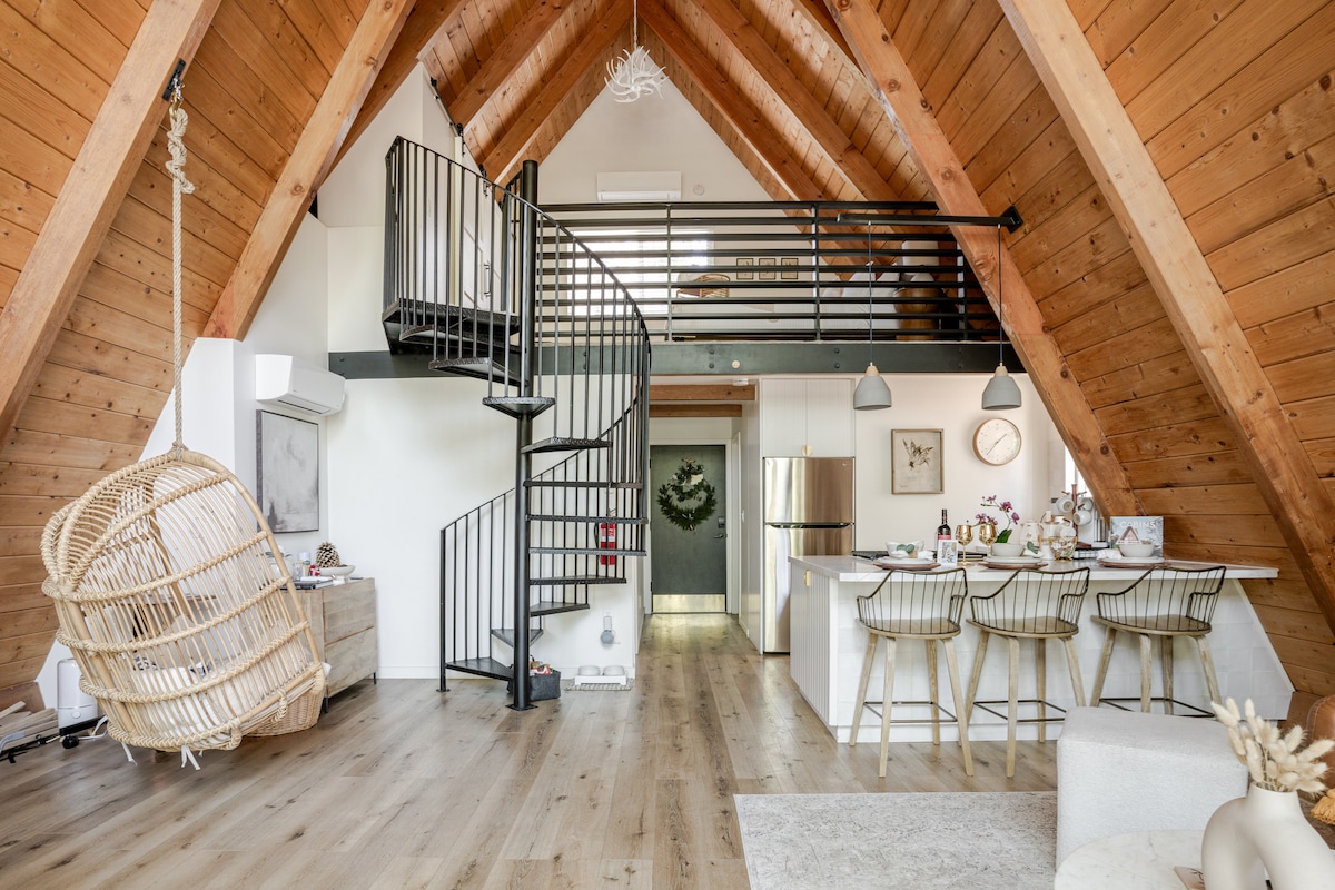 An A-frame cabin interior showcases a high ceiling with wooden beams, creating a spacious atmosphere. A spiral staircase leads to the loft area, while a dining bar with stylish stools connects the kitchen to the living space. A hanging chair adds a touch of comfort.