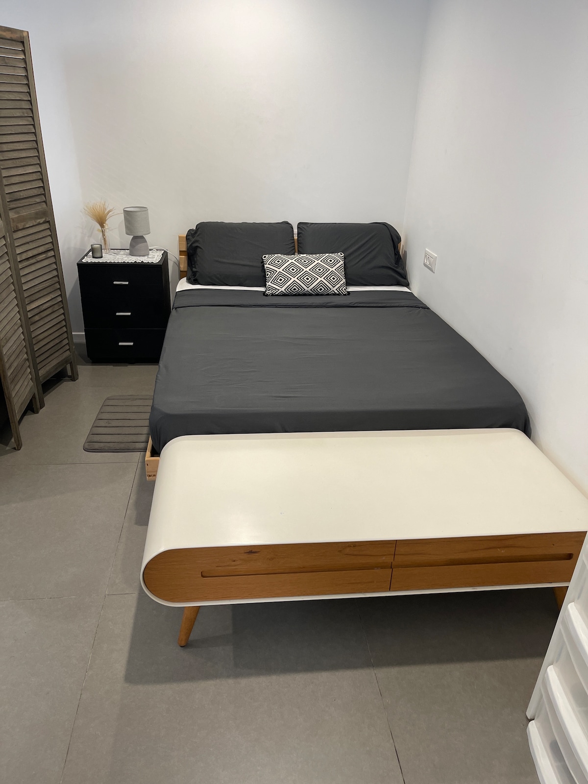 A simple sleeping area features a spacious bed with dark linens and decorative pillows. A mid-century modern table is placed at the foot of the bed, complementing a nearby nightstand. Soft lighting illuminates the room, enhancing the minimalist design.